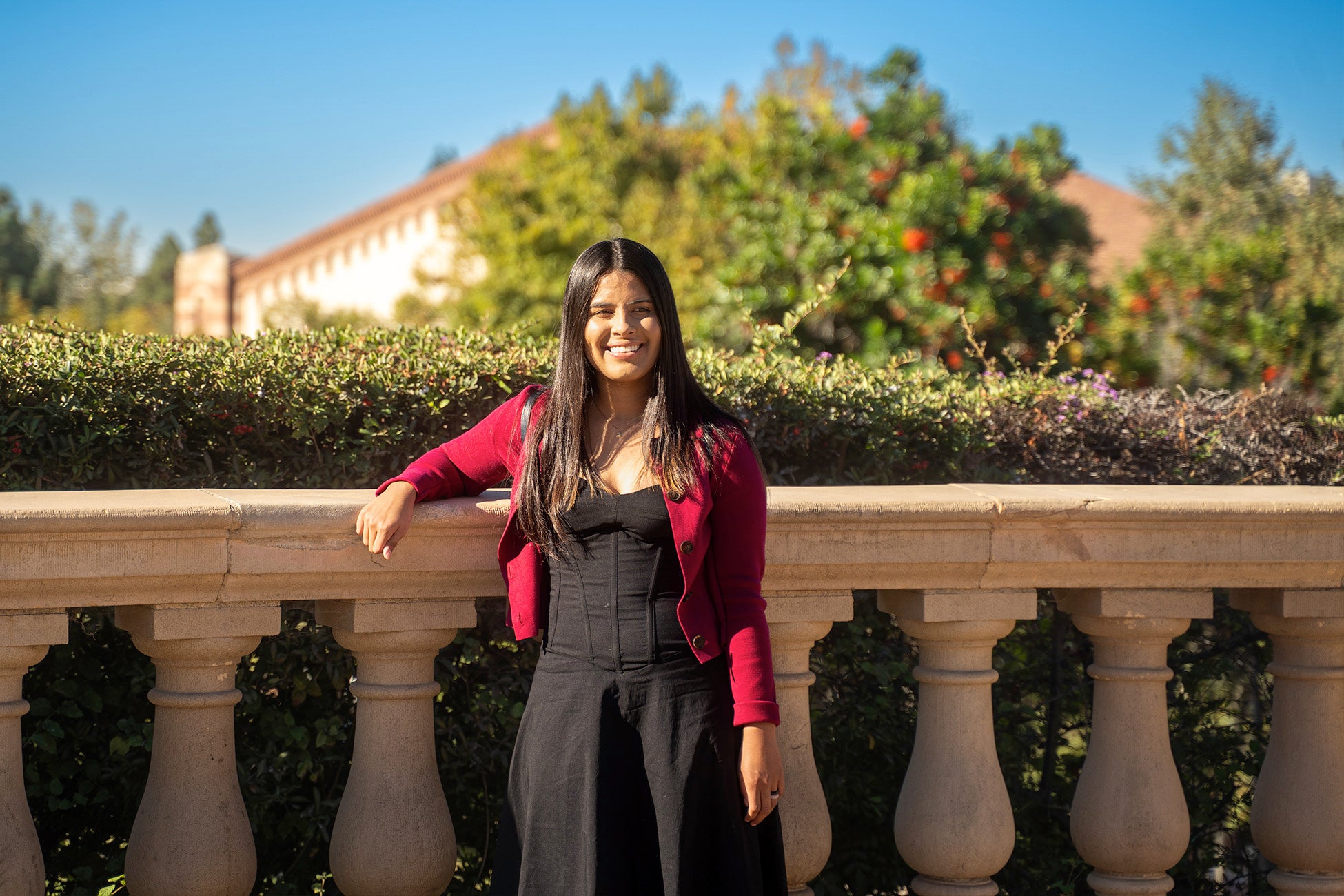 A woman with long dark hair stands smiling outdoors, leaning against a stone balustrade. She wears a black dress with a red cardigan, with greenery, flowering bushes, and a campus building blurred in the sunny background.